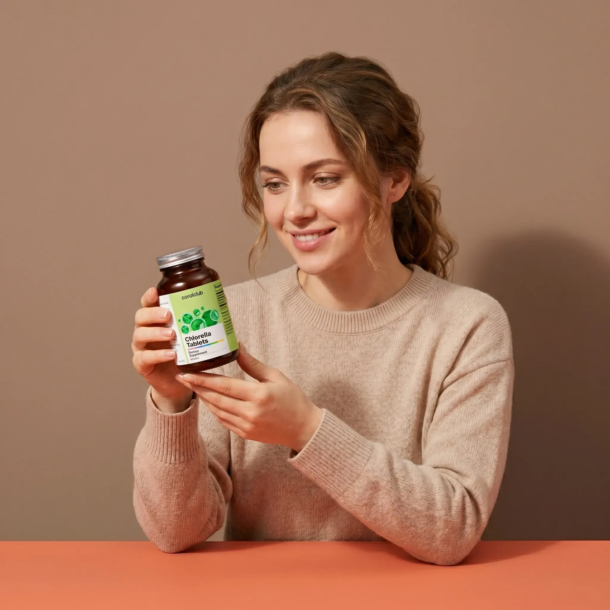 Woman holding a bottle of Chlorella Tablets, smiling, in a cozy indoor setting.