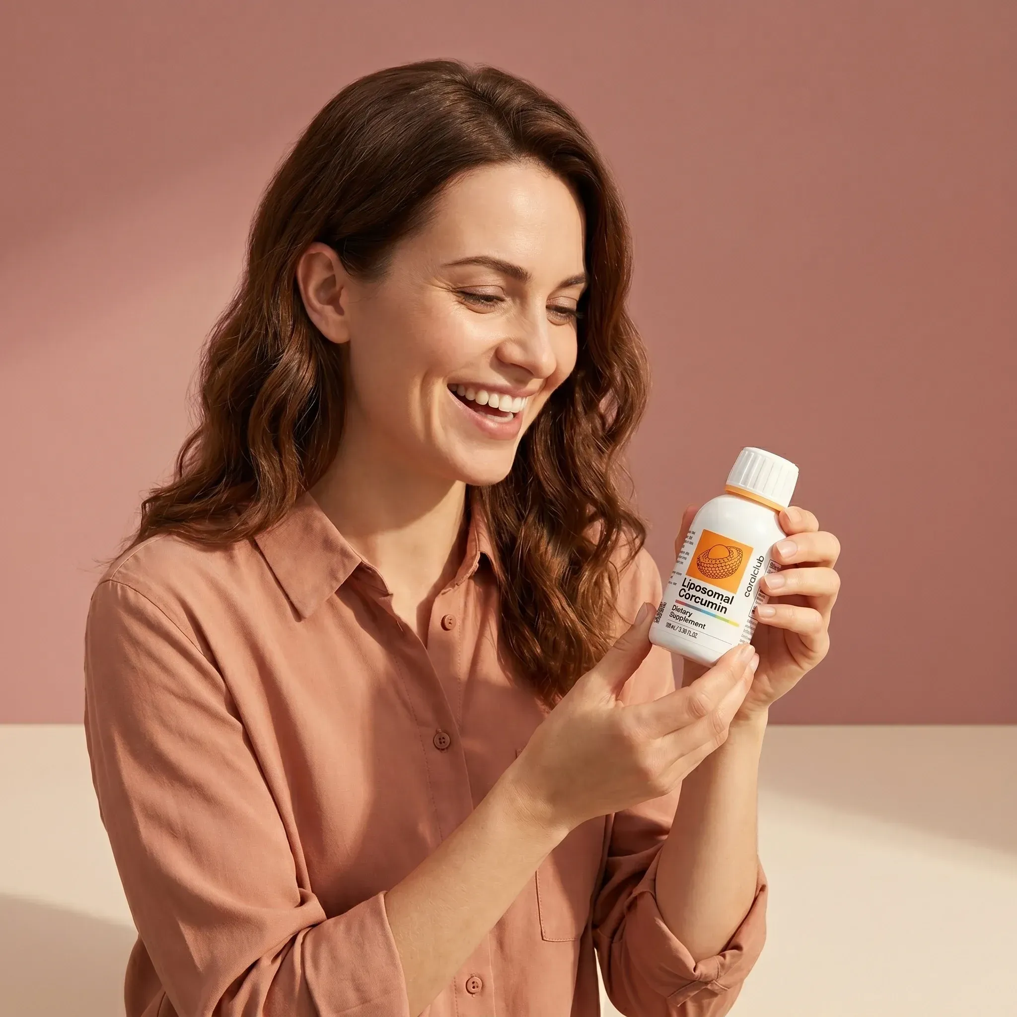 Woman smiling while holding a bottle of Liposomal Curcumin against a soft pink background.
